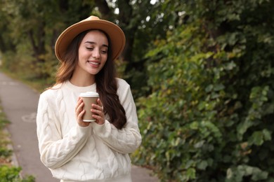 Photo of Beautiful young woman in stylish warm sweater holding paper cup of coffee outdoors, space for text