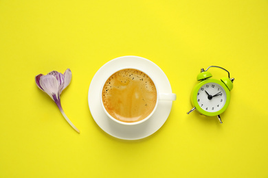 Photo of Delicious morning coffee, alarm clock and flower on yellow background, flat lay