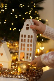 Woman holding decorative house near Christmas decorations on window sill indoors, closeup