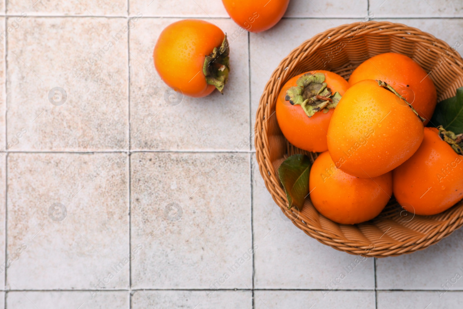 Photo of Delicious ripe juicy persimmons in wicker basket on tiled surface, flat lay. Space for text