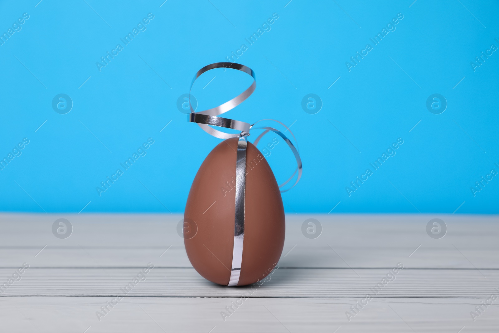 Delicious chocolate egg with silver ribbon on white wooden table against light blue background Photo of Delicious chocolate egg with silver ribbon on white wooden table against light blue background