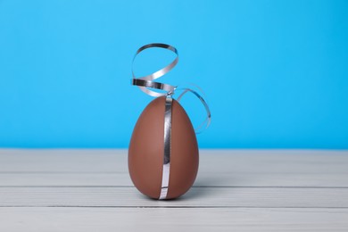 Photo of Delicious chocolate egg with silver ribbon on white wooden table against light blue background