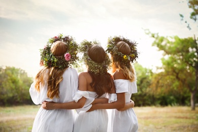 Young women wearing wreaths made of beautiful flowers outdoors on sunny day, back view