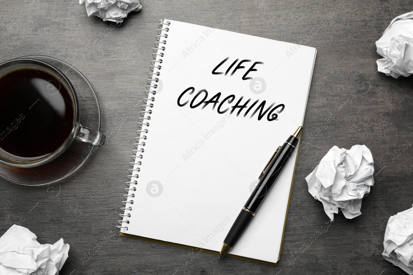 Image of Phrase Life Coaching written in notebook, cup of coffee, crumpled paper and pen on grey table, flat lay