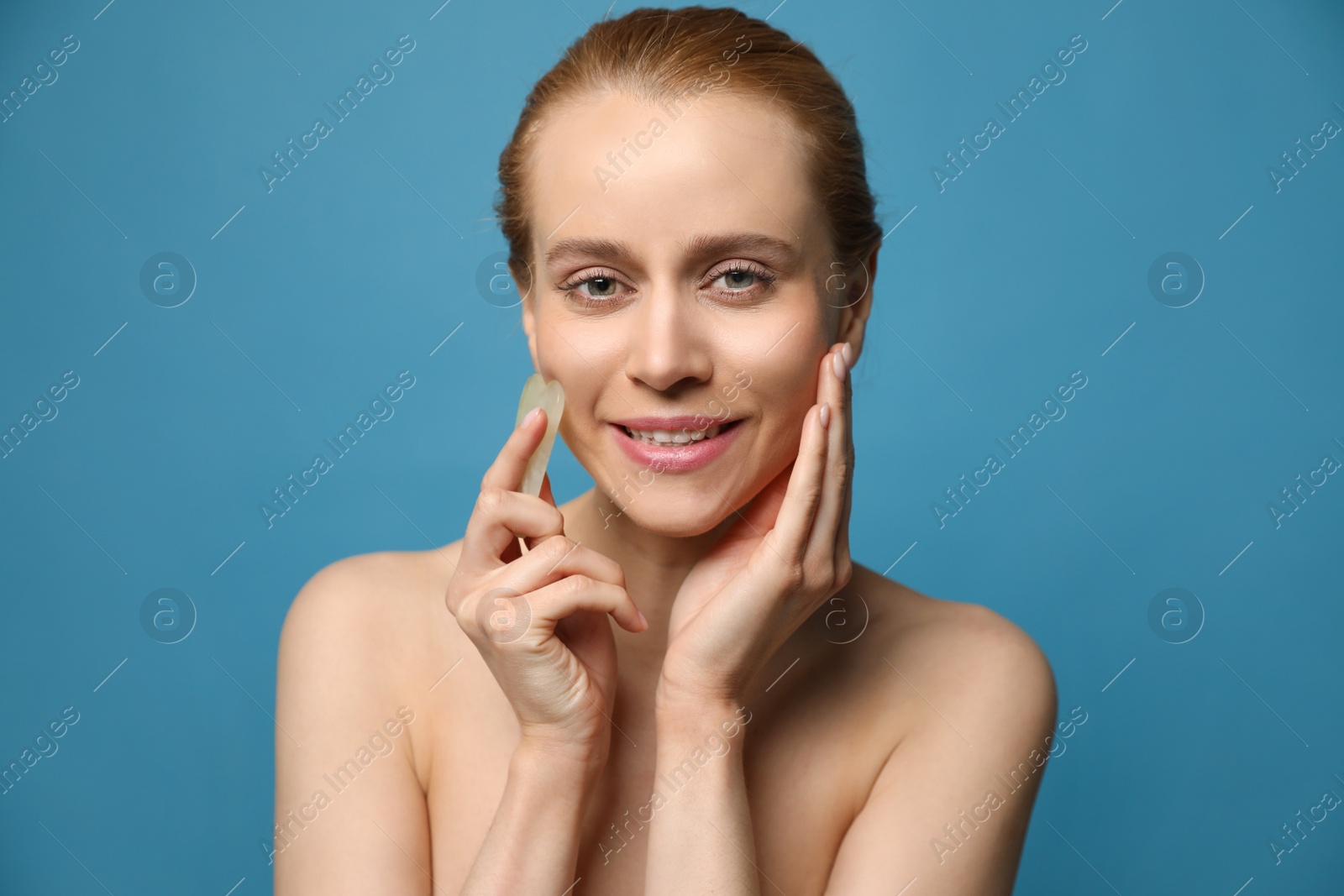 Photo of Beautiful young woman doing facial massage with gua sha tool on blue background