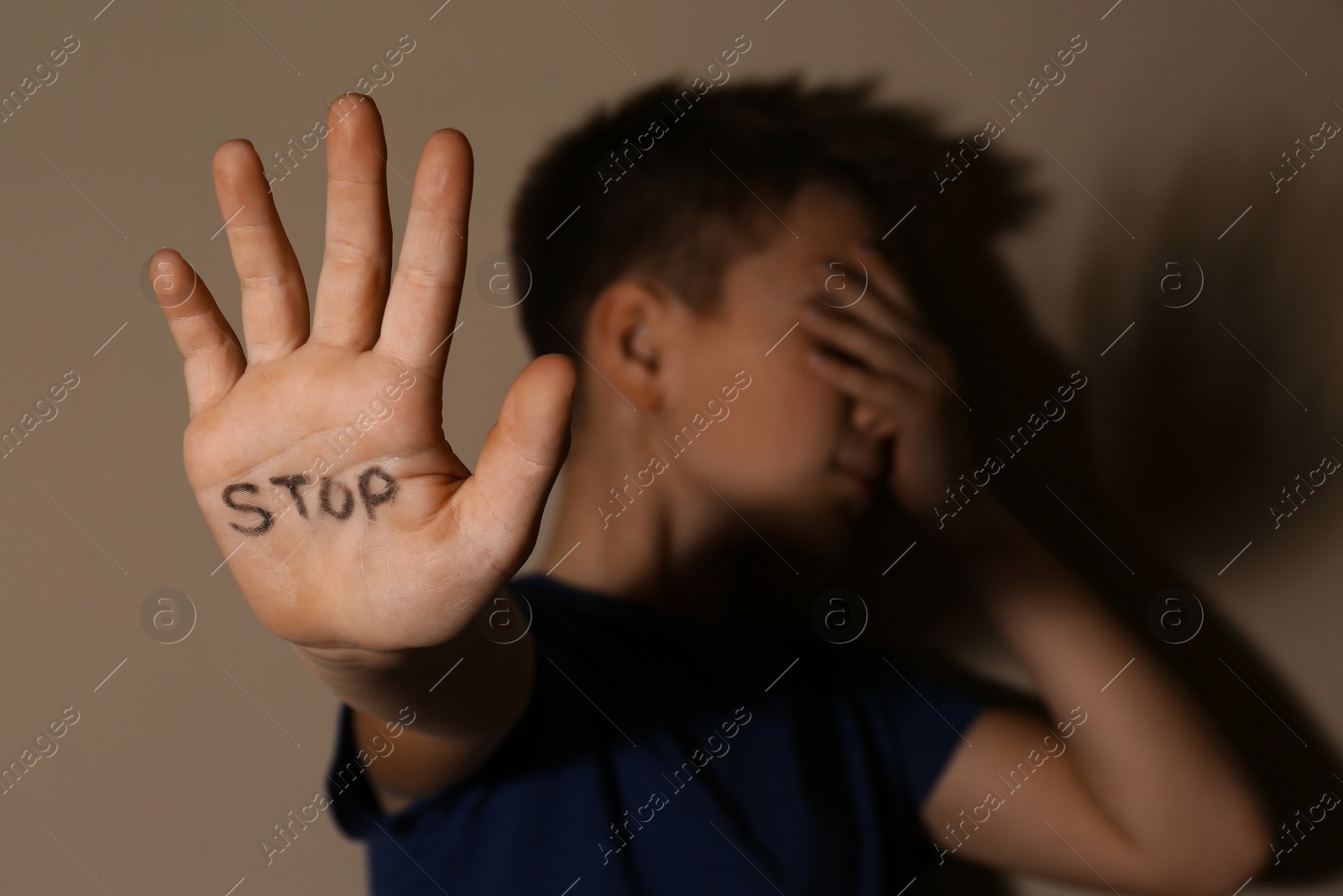 Photo of Abused little boy showing palm with word STOP near beige wall, focus on hand. Domestic violence concept