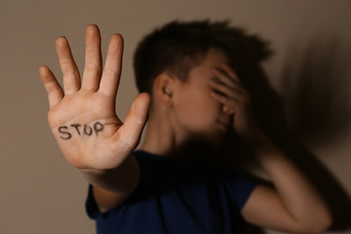 Photo of Abused little boy showing palm with word STOP near beige wall, focus on hand. Domestic violence concept