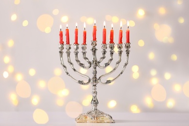 Photo of Silver menorah with burning candles against light grey background and blurred festive lights. Hanukkah celebration