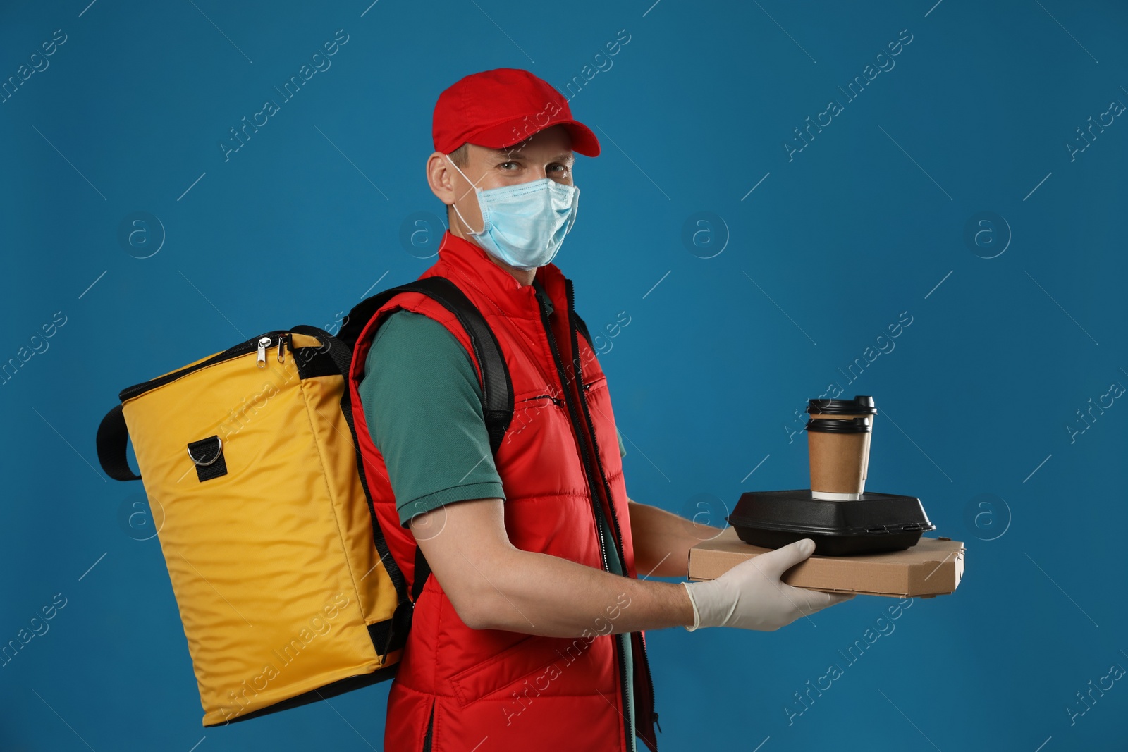 Photo of Courier in protective mask and gloves holding order on blue background. Food delivery service during coronavirus quarantine