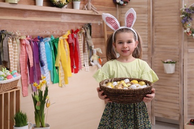 Photo of Happy little girl with bunny ears and wicker basket full of Easter eggs indoors