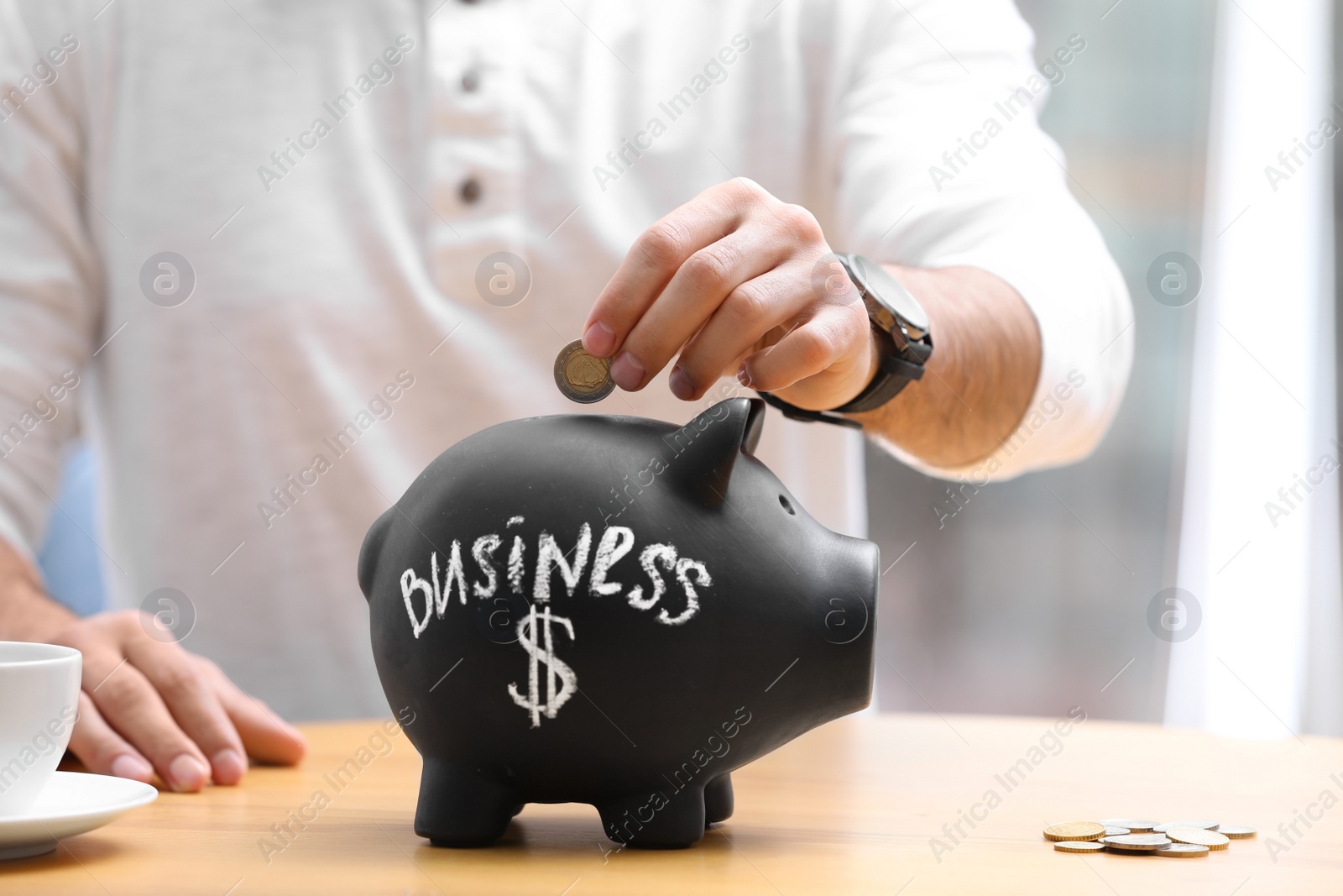 Photo of Man putting money into piggy bank with word BUSINESS at table, closeup
