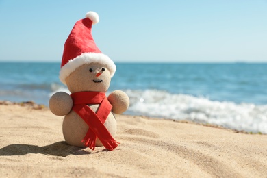 Photo of Snowman made of sand with Santa hat and scarf on beach near sea, space for text. Christmas vacation