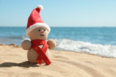 Snowman made of sand with Santa hat and scarf on beach near sea, space for text. Christmas vacation