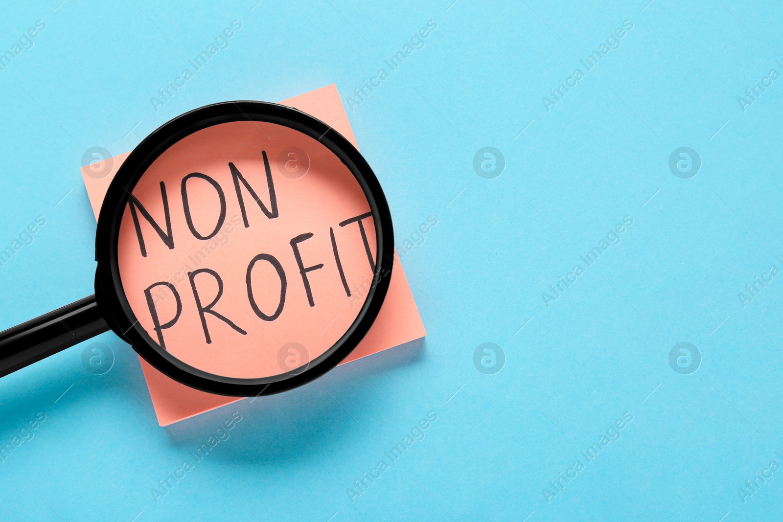 Photo of Paper note with phrase Non Profit under magnifying glass on light blue background, top view. Space for text