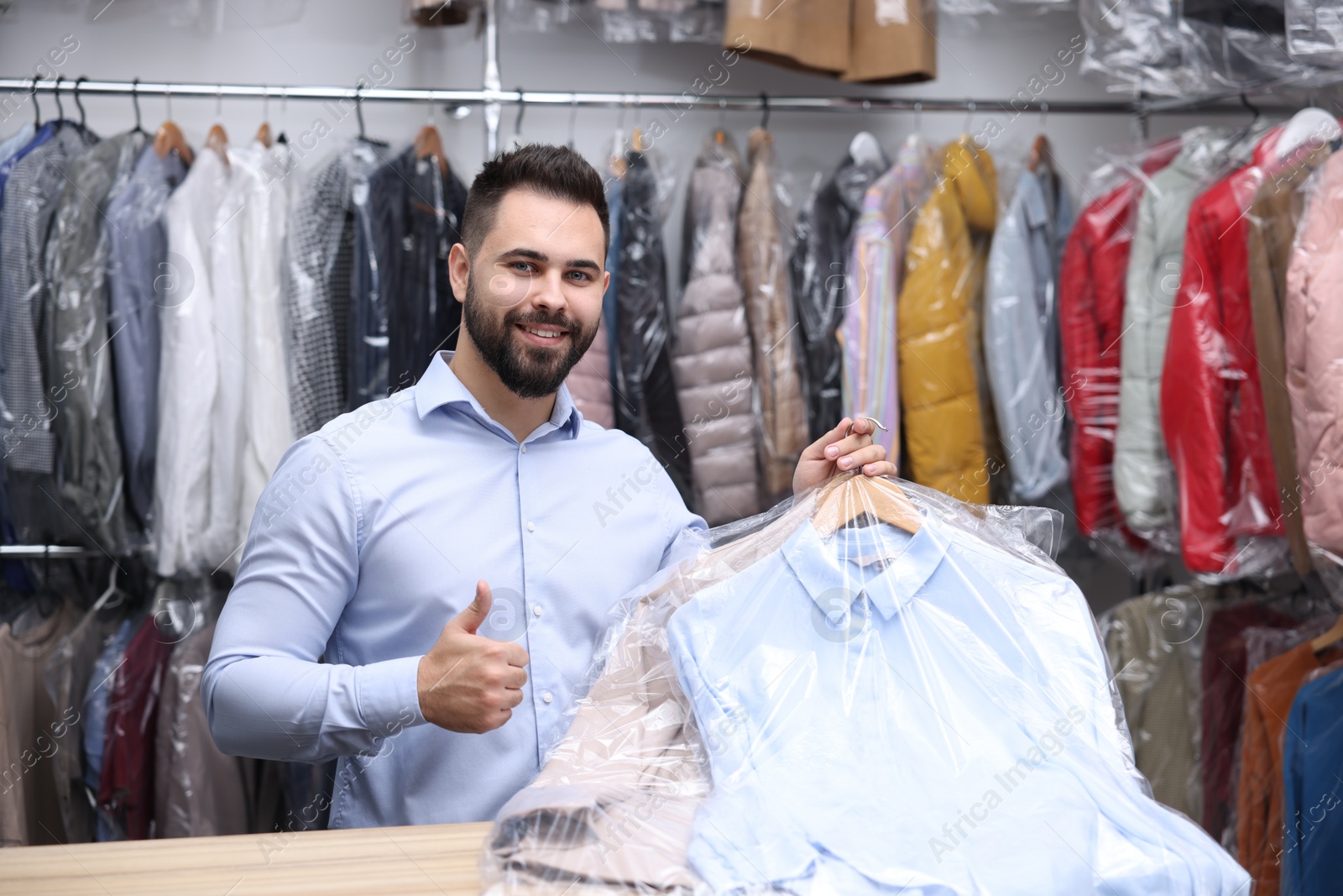 Photo of Dry-cleaning service. Happy worker holding hangers with clothes in plastic bags and showing thumb up at counter indoors