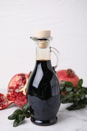 Photo of Tasty pomegranate sauce in bottle, fruits and branches on white marble table, closeup