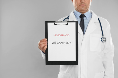 Doctor holding clipboard with words HEMORRHOID WE CAN HELP on light grey background, closeup