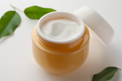 Photo of Jar of face cream and green leaves on white background, closeup