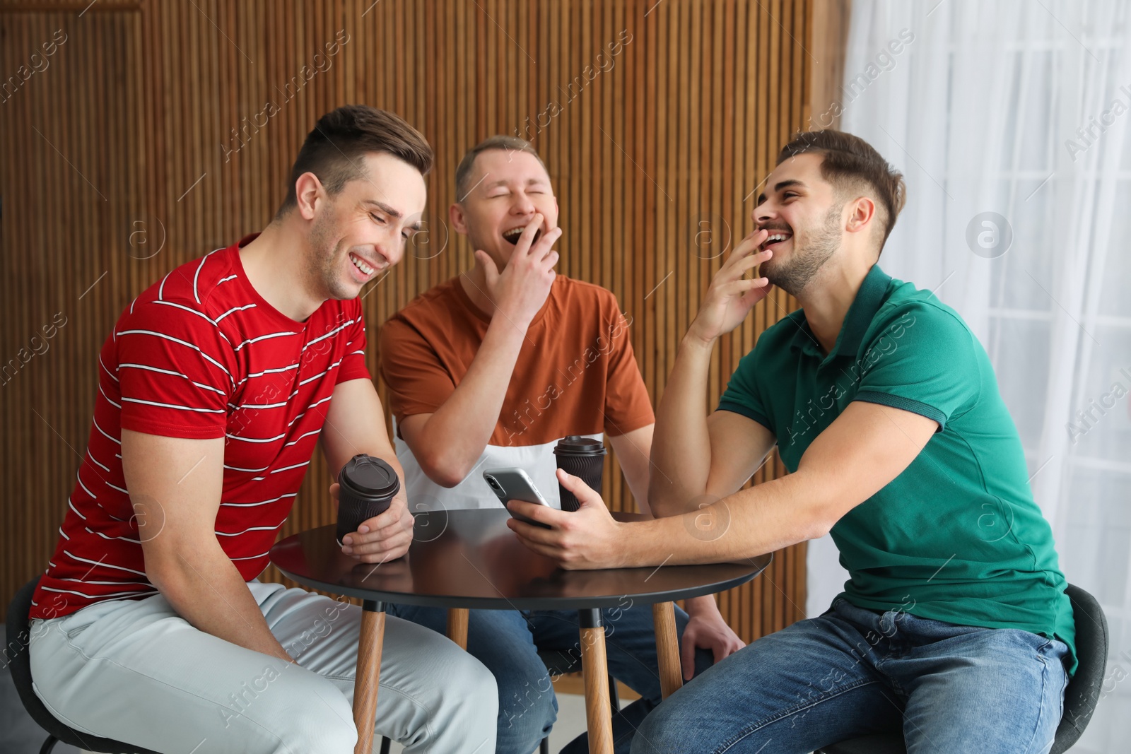 Photo of Men with cups of coffee and smartphone laughing together at table indoors