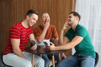 Photo of Men with cups of coffee and smartphone laughing together at table indoors