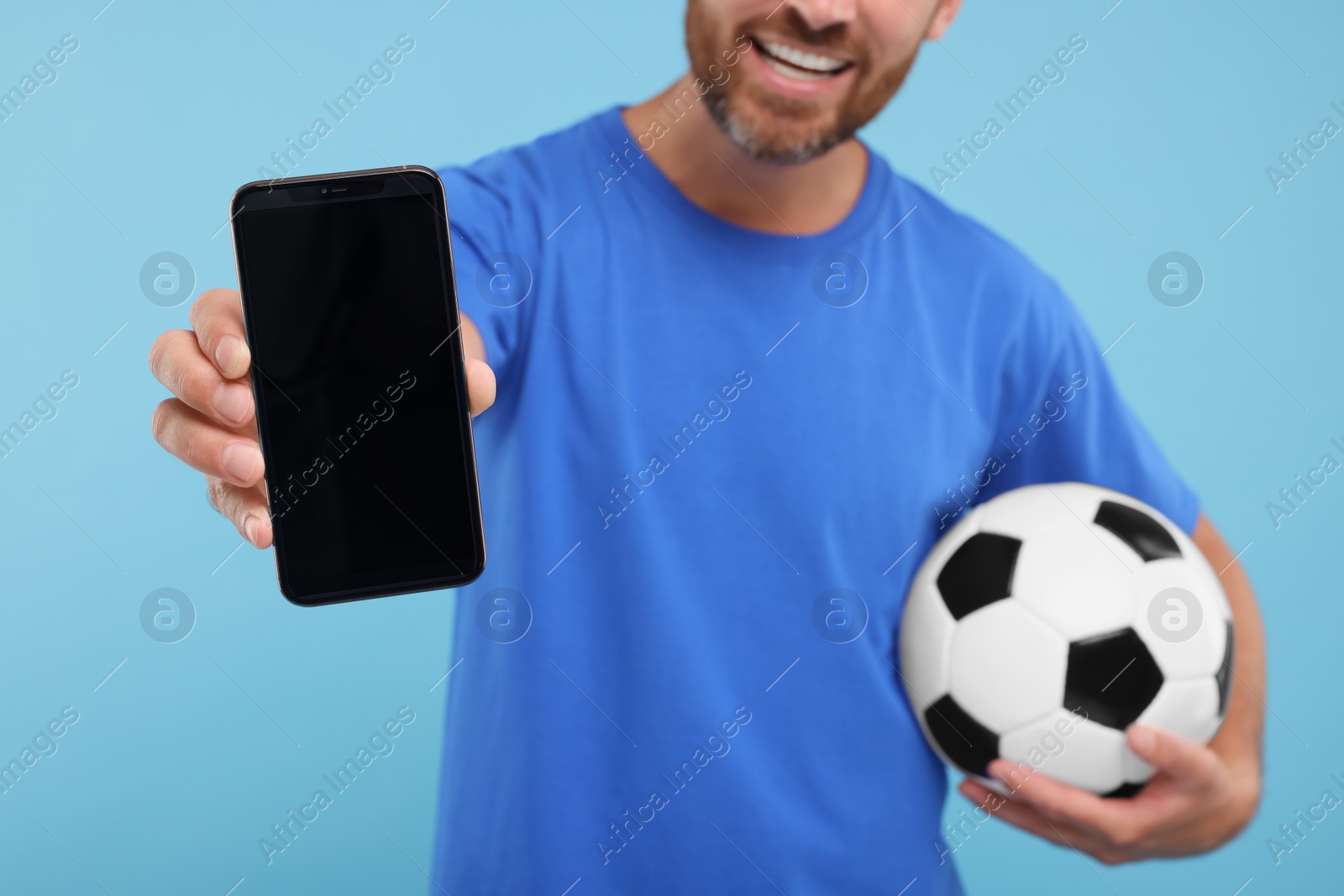 Photo of Happy sports fan with soccer ball and smartphone on light blue background, closeup