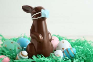 Photo of Chocolate bunny with protective mask and eggs against white background, closeup. Easter holiday during 
COVID-19 quarantine