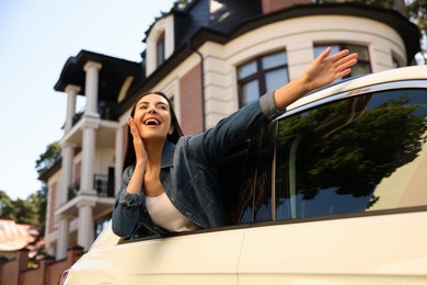 Happy young woman leaning out of window while enjoying car trip on city street, low angle view