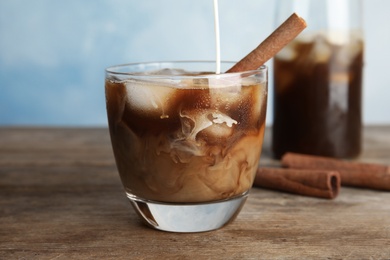 Pouring milk into glass of coffee with ice cubes on table against color background