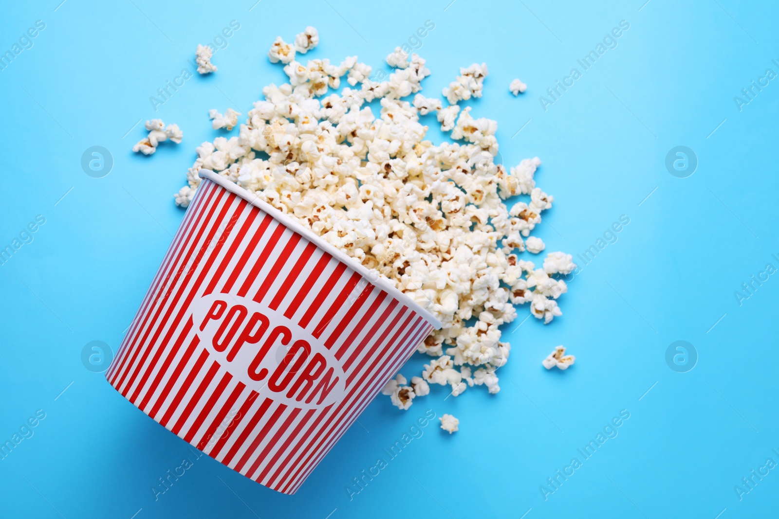 Photo of Overturned paper bucket with delicious popcorn on light blue background, flat lay