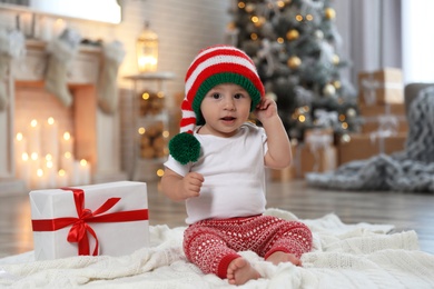 Image of Cute little baby with elf hat near Christmas gift on floor at home