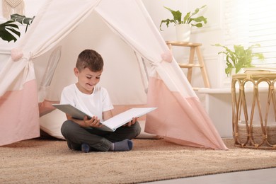Photo of Cute little boy reading book near toy wigwam at home