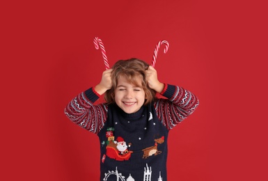 Photo of Cute little girl in Christmas sweater holding sweet candy canes near head against red background