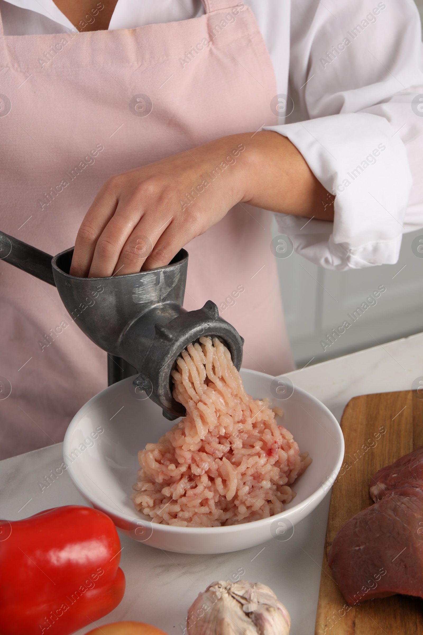 Photo of Woman making chicken mince with metal meat grinder at white table in kitchen, closeup