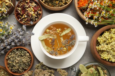 Photo of Cup of aromatic freshly brewed tea surrounded by different dry herbs on grey table, flat lay