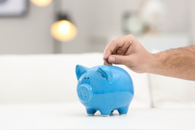 Photo of Young man putting money into piggy bank at white table indoors, closeup