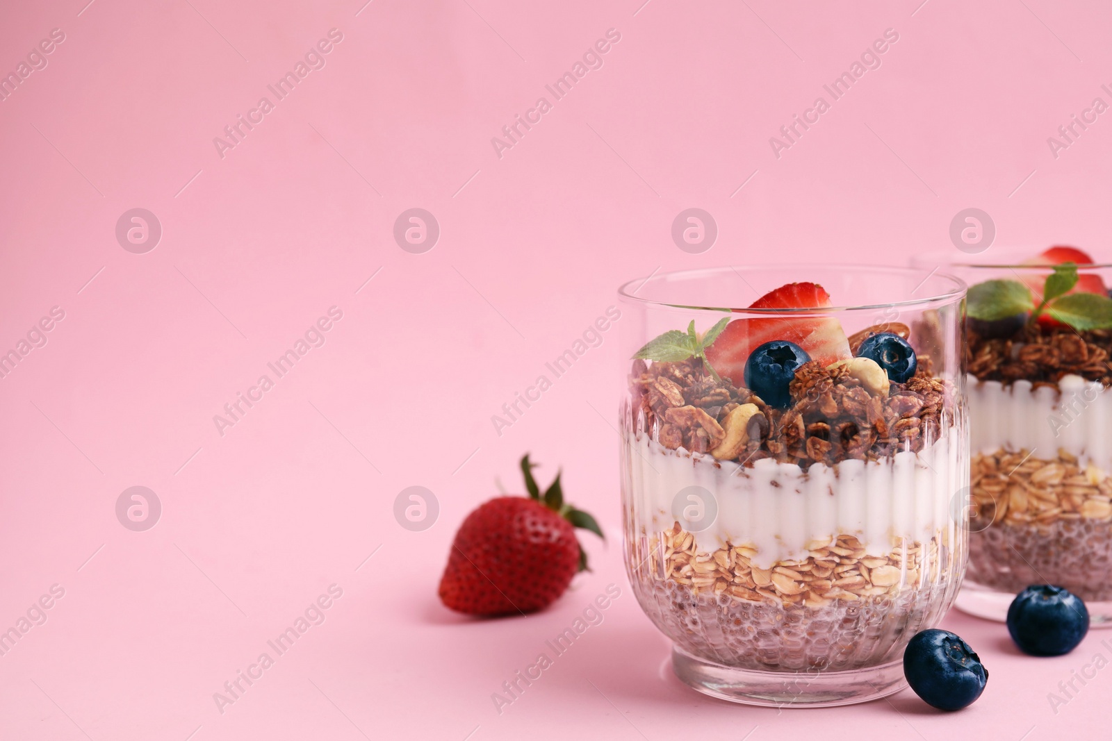 Photo of Tasty granola with berries, nuts, yogurt and chia seeds in glasses on pink background, space for text