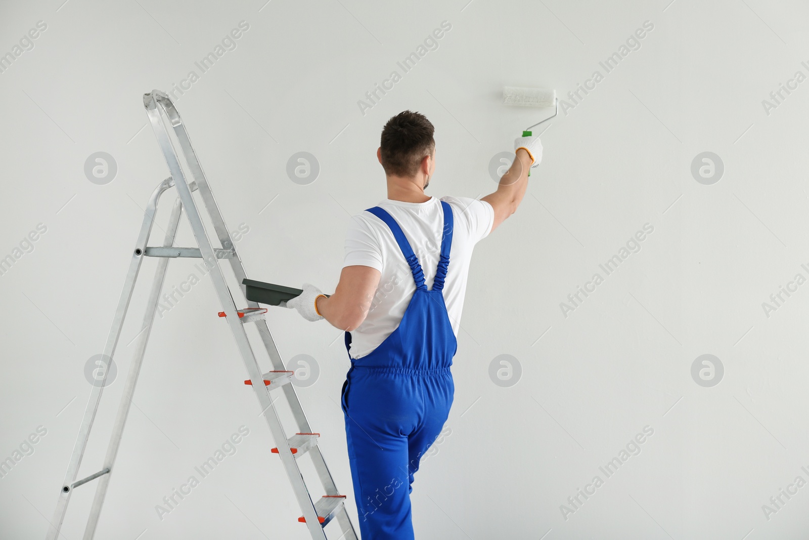 Photo of Man painting wall with white dye indoors, back view