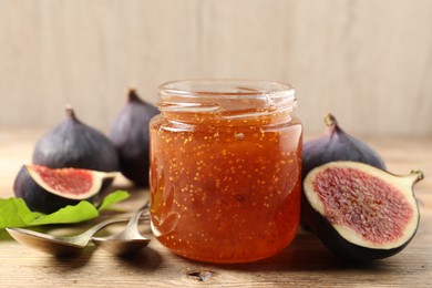 Glass jar with tasty sweet jam, spoons and fresh figs on wooden table, closeup