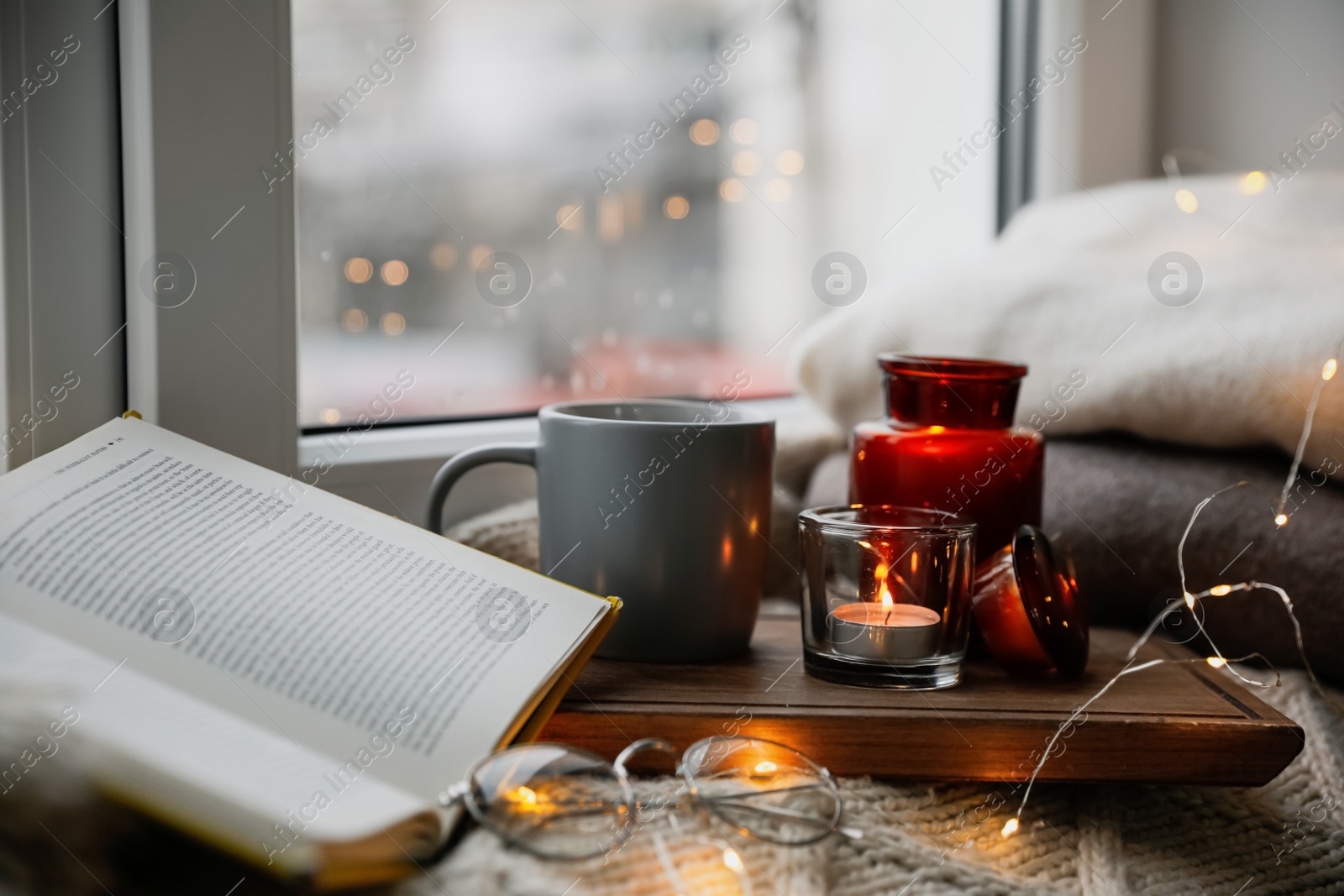 Photo of Cup of hot drink, open book and Christmas lights on knitted blanket near window. Cozy season