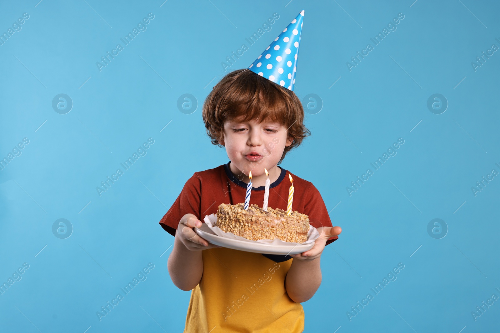 Photo of Birthday celebration. Cute little boy in party hat blowing candles on tasty cake against light blue background