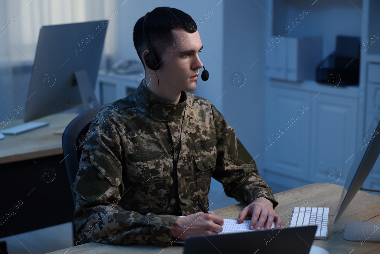 Photo of Military service. Young soldier in headphones working at wooden table in office at night