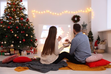 Photo of Couple eating snacks and watching Christmas movie via video projector in cosy room, back view. Winter holidays atmosphere
