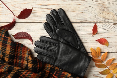 Photo of Stylish black leather gloves, scarf and dry leaves on white wooden table, flat lay