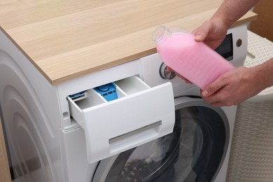 Photo of Man pouring fabric softener from bottle into washing machine indoors, closeup