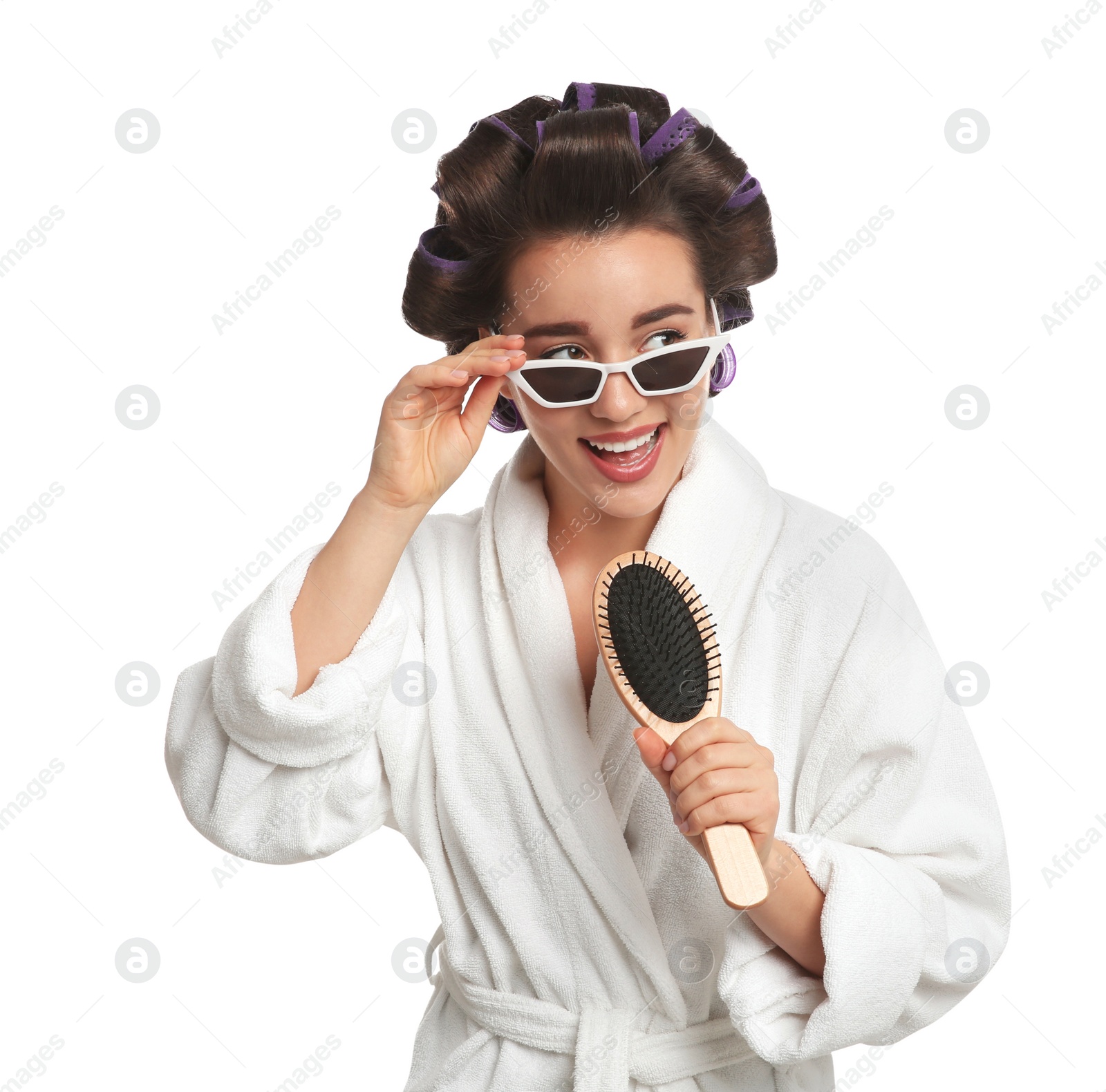 Photo of Happy young woman in bathrobe with hair curlers and sunglasses singing into hairbrush on white background