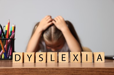 Photo of Little girl covering head with hands and cubes with word Dyslexia at wooden table, selective focus