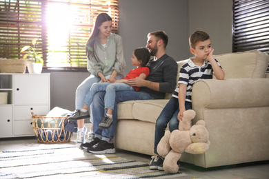 Photo of Unhappy little boy feeling jealous while parents spending time with his sister at home