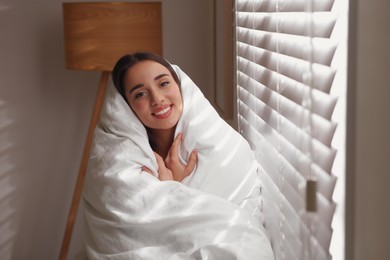 Beautiful young woman wrapped with soft blanket near window at home