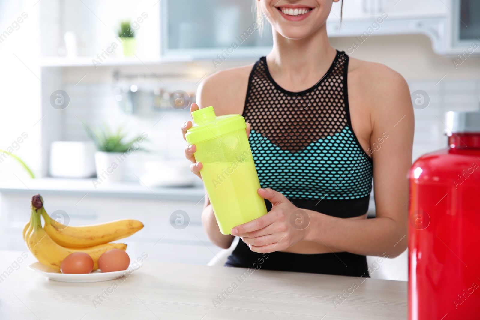 Photo of Young woman holding bottle of protein shake at table with ingredients in kitchen, closeup. Space for text