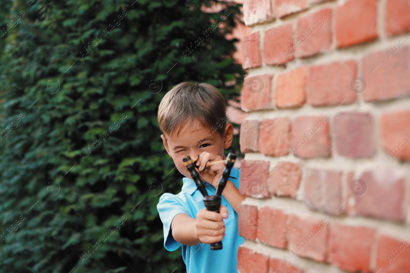 Photo of Little boy playing with slingshot near brick wall outdoors